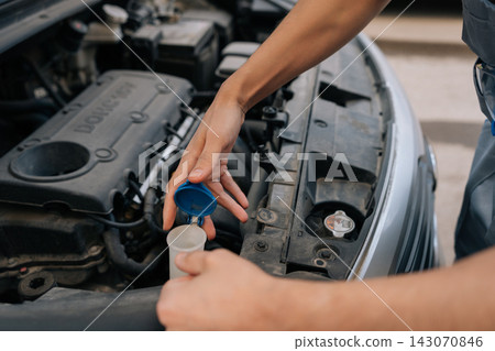 Close-up shot of auto mechanic performing vehicle maintenance. Unrecognizable person opens blue cap of washer fluid reservoir to check liquid level in engine compartment. Focus on hands and action. Close-up shot of auto mechanic performing vehicle maintenance. Unrecognizable person opens blue cap of washer fluid reservoir to check liquid level in engine compartment. Focus on hands and action. 143070846