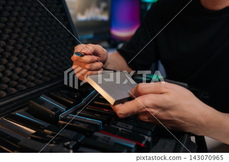 Closeup of technician hands holding portable SSD hard drive and USB cable. Man prepares to connect device for data backup or transfer. Dark tech setting with equipment case full of drives. Closeup of technician hands holding portable SSD hard drive and USB cable. Man prepares to connect device for data backup or transfer. Dark tech setting with equipment case full of drives. 143070965
