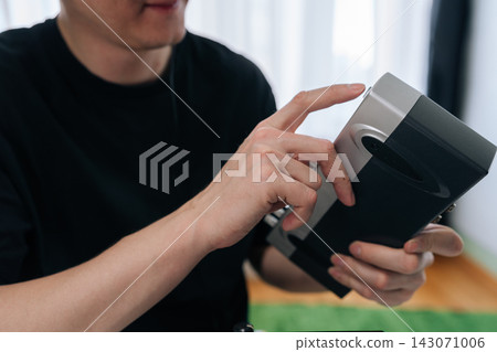 Man in black t shirt holding and touching portable smart device at home, close-up of hands interacting with touchscreen, showcasing modern connectivity and everyday tech use. Man in black t shirt holding and touching portable smart device at home, close-up of hands interacting with touchscreen, showcasing modern connectivity and everyday tech use. 143071006