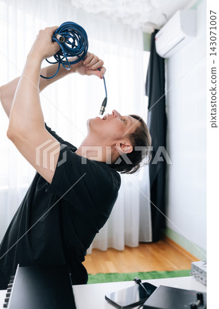 Funny musician or artist in black t-shirt pretends to sing into or eat blue audio cable. Bizarre concept of creativity, sound, or hunger for music in bright home studio setting. Funny musician or artist in black t-shirt pretends to sing into or eat blue audio cable. Bizarre concept of creativity, sound, or hunger for music in bright home studio setting. 143071007