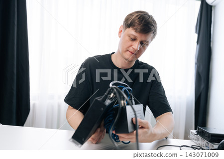 Focused man in black t-shirt sitting at white table, examining and assembling black electronic equipment with tangled blue wires. Bright background with natural light from window. Focused man in black t-shirt sitting at white table, examining and assembling black electronic equipment with tangled blue wires. Bright background with natural light from window. 143071009