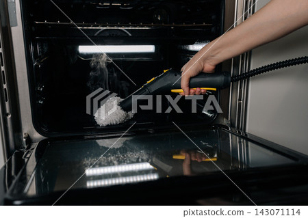 Close-up of hand cleaning oven interior with steam cleaner, applying high-pressure vapor to remove grime and sanitize surfaces for thorough home kitchen maintenance and hygiene. Close-up of hand cleaning oven interior with steam cleaner, applying high-pressure vapor to remove grime and sanitize surfaces for thorough home kitchen maintenance and hygiene. 143071114