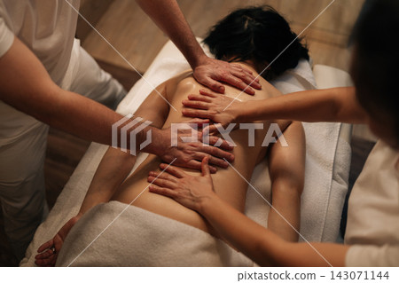 High-angle view of male and female professional masseurs performing relaxing four hand massage on woman back, using warm oil in dark wellness center for deep muscle relief, rejuvenation body care. 143071144
