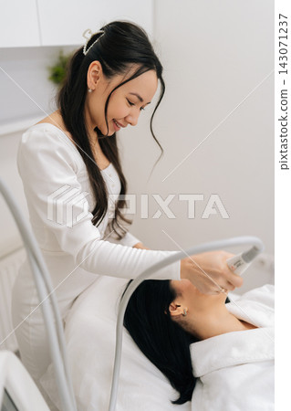 Smiling positive Asian beautician performing innovative microneedle rf lifting treatment on female patient face, utilizing advanced radiofrequency technology for skin tightening and rejuvenation. Smiling positive Asian beautician performing innovative microneedle rf lifting treatment on female patient face, utilizing advanced radiofrequency technology for skin tightening and rejuvenation. 143071237
