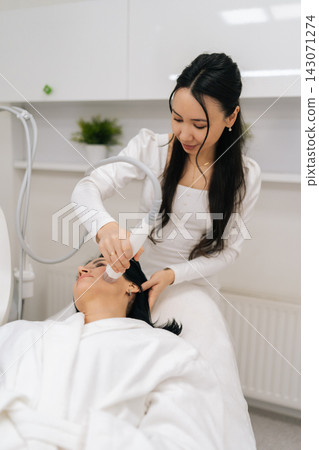 Vertical portrait of young Asian cosmetologist performing fractional microneedle rf lifting procedure on mature female patient face in modern beauty clinic for skin rejuvenation and wrinkle reduction. Vertical portrait of young Asian cosmetologist performing fractional microneedle rf lifting procedure on mature female patient face in modern beauty clinic for skin rejuvenation and wrinkle reduction. 143071274