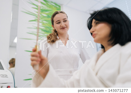Young female cosmetologist consulting with middle-aged woman client in modern salon, examining reflection in hand mirror after facial treatment, discussing results and care plans. Young female cosmetologist consulting with middle-aged woman client in modern salon, examining reflection in hand mirror after facial treatment, discussing results and care plans. 143071277