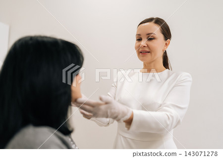 Portrait of professional female cosmetologist wearing gloves talking to mature woman patient in chair and examining facial skin before acosmetic procedure in modern aesthetic clinic. Portrait of professional female cosmetologist wearing gloves talking to mature woman patient in chair and examining facial skin before acosmetic procedure in modern aesthetic clinic. 143071278