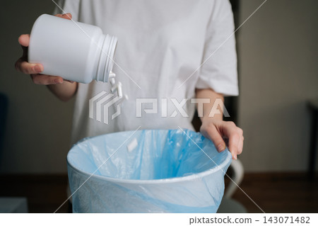 Closeup of person discarding expired pills and capsules from white bottle into blue-bagged trash can, illustrating responsible medication disposal, household safety and environmental care. Closeup of person discarding expired pills and capsules from white bottle into blue-bagged trash can, illustrating responsible medication disposal, household safety and environmental care. 143071482