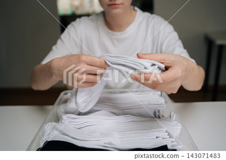 Woman neatly folding clean white socks into clear storage box, illustrating efficient laundry organization, tidy home routine, minimalist closet storage for everyday garments, close-up cropped shot. Woman neatly folding clean white socks into clear storage box, illustrating efficient laundry organization, tidy home routine, minimalist closet storage for everyday garments, close-up cropped shot. 143071483