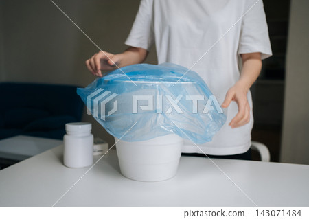 Person lining white bin with blue plastic garbage bag next to medicine bottle on white table, representing responsible disposal of unwanted medication, withdrawal, new beginning. Person lining white bin with blue plastic garbage bag next to medicine bottle on white table, representing responsible disposal of unwanted medication, withdrawal, new beginning. 143071484