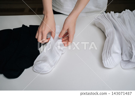 Close-up of hands folding pair of freshly laundered white socks on clean white surface, preparing for organized storage alongside stack of other folded white socks and pile of black socks. Close-up of hands folding pair of freshly laundered white socks on clean white surface, preparing for organized storage alongside stack of other folded white socks and pile of black socks. 143071487