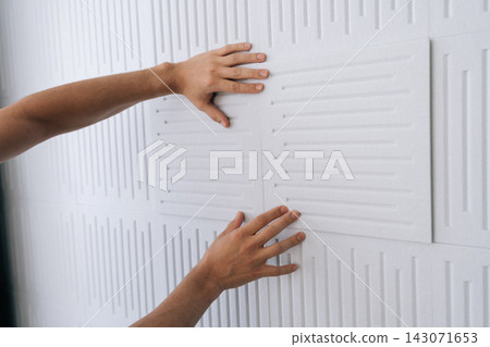 Hands installing soundproofing acoustic panels on a white studio wall, creating a quiet environment for sound dampening and noise reduction in a recording space. 143071653