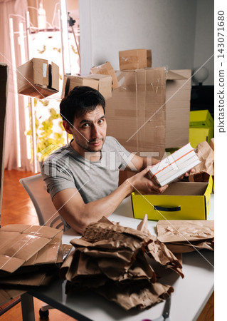 Portrait of man looking at camera packing books into yellow box at table. Messy room filled with cardboard containers and packing paper. Concept of moving, relocating, organizing home. Portrait of man looking at camera packing books into yellow box at table. Messy room filled with cardboard containers and packing paper. Concept of moving, relocating, organizing home. 143071680
