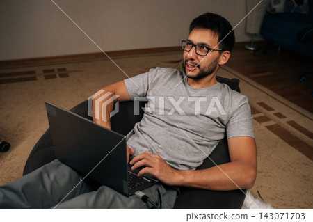 Portrait of young bearded man wearing glasses and casual clothing, sitting comfortably in bean bag chair and looking away from laptop while engaging in online work from comfy home environment. Portrait of young bearded man wearing glasses and casual clothing, sitting comfortably in bean bag chair and looking away from laptop while engaging in online work from comfy home environment. 143071703