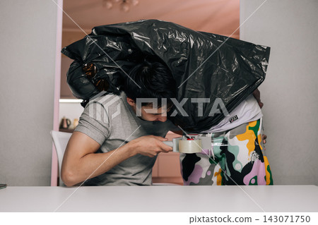 Young man using packing tape to humorously wrap second person inside large black garbage bag, creating playful and surprising moment for prank. 143071750