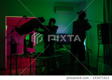 Production crew preparing indoor film set with lighting and boom mic, adjusting equipment under vibrant green and pink illumination for video production and content creation. Production crew preparing indoor film set with lighting and boom mic, adjusting equipment under vibrant green and pink illumination for video production and content creation. 143071822