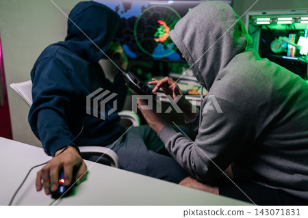 Two unrecognizable people in hooded sweatshirts sit at desk in dark room. They work on computer, focused on screen with world map and code. Green light creates mysterious, tense atmosphere. Two unrecognizable people in hooded sweatshirts sit at desk in dark room. They work on computer, focused on screen with world map and code. Green light creates mysterious, tense atmosphere. 143071831