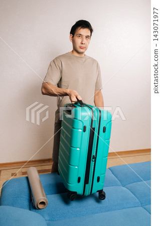 Vertical portrait of traveler male holding suitcase, preparing for travel while considering luggage protection, with roll of clear plastic wrap on sofa, emphasizing vacation planning and anticipation. Vertical portrait of traveler male holding suitcase, preparing for travel while considering luggage protection, with roll of clear plastic wrap on sofa, emphasizing vacation planning and anticipation. 143071977