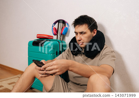 Male traveler sitting on floor with neck pillow and smartphone, having a delay, waiting for departure, feeling impatient while checking updates on mobile device during trip. Male traveler sitting on floor with neck pillow and smartphone, having a delay, waiting for departure, feeling impatient while checking updates on mobile device during trip. 143071999
