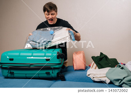 Man expressing surprise while packing clothes in vacuum storage bags for efficient luggage space, preparing for travel or vacation. Man expressing surprise while packing clothes in vacuum storage bags for efficient luggage space, preparing for travel or vacation. 143072002