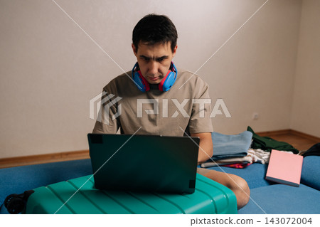 Young man preparing for travel, working on his laptop placed on a teal luggage, with neatly folded clothes stacked beside him on a couch, organizing logistics for his vacation. Young man preparing for travel, working on his laptop placed on a teal luggage, with neatly folded clothes stacked beside him on a couch, organizing logistics for his vacation. 143072004