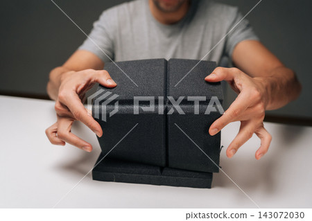 Close-up of man holding black acoustic foam panel, examining sound absorbing material for noise control, soundproofing, insulation in dark recording studio or home environment. Close-up of man holding black acoustic foam panel, examining sound absorbing material for noise control, soundproofing, insulation in dark recording studio or home environment. 143072030