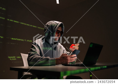 Dangerous hooded hacker male in sunglasses using smartphone and holding red credit card, surrounded by green code visuals suggesting cybercrime, fraud and identity theft. Dangerous hooded hacker male in sunglasses using smartphone and holding red credit card, surrounded by green code visuals suggesting cybercrime, fraud and identity theft. 143072054