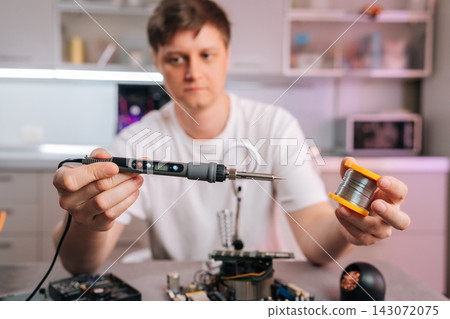 Man holding modern soldering iron and spool of solder wire, preparing to repair or assemble an electronic circuit board, concept of process of technical maintenance and diy electronics. Man holding modern soldering iron and spool of solder wire, preparing to repair or assemble an electronic circuit board, concept of process of technical maintenance and diy electronics. 143072075