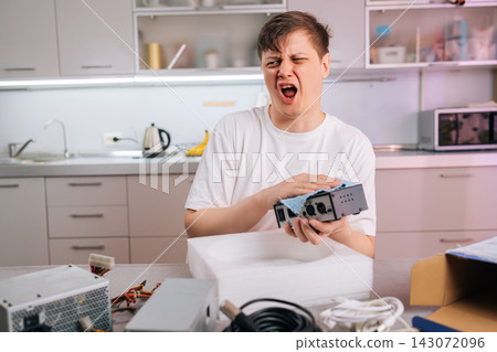 Man makes disgusted face cleaning dusty electronic part with blue cloth. He sneezes from dust allergy or frustrated with tech repair. Messy foreground with computer components in modern kitchen. Man makes disgusted face cleaning dusty electronic part with blue cloth. He sneezes from dust allergy or frustrated with tech repair. Messy foreground with computer components in modern kitchen. 143072096