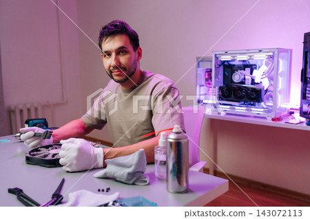 Smiling technician cleaning dusty computer fan with brush. Man in gloves at desk performs PC maintenance. Workshop has custom computer with purple lighting. Concept of hardware repair, service, hobby. Smiling technician cleaning dusty computer fan with brush. Man in gloves at desk performs PC maintenance. Workshop has custom computer with purple lighting. Concept of hardware repair, service, hobby. 143072113