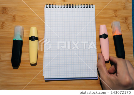 Flat lay of open grid notebook with colorful markers. Hand holding pink highlighter on wooden background. Top down view of office supplies for studying, planning or creative work. Blank copy space. Flat lay of open grid notebook with colorful markers. Hand holding pink highlighter on wooden background. Top down view of office supplies for studying, planning or creative work. Blank copy space. 143072170