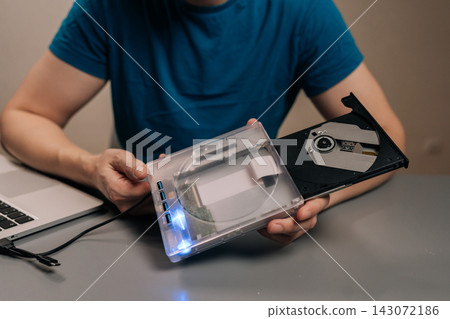 Close-up hand of person holding transparent external optical drive with open disc tray connected to laptop on desk, demonstrating portable dvd cd reading, writing and data transfer for work or home. Close-up hand of person holding transparent external optical drive with open disc tray connected to laptop on desk, demonstrating portable dvd cd reading, writing and data transfer for work or home. 143072186