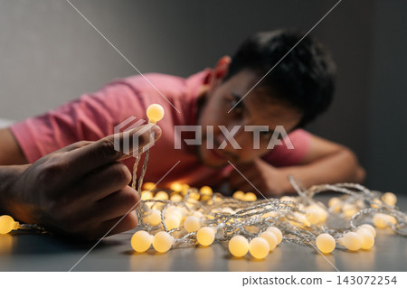 Happy young man holding glowing decorative light bulb from string garland, representing anticipation of festive occasions and bringing warmth to interior decor. Happy young man holding glowing decorative light bulb from string garland, representing anticipation of festive occasions and bringing warmth to interior decor. 143072254