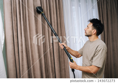 Portrait of young bearded man professionally steam cleaning curtains in modern home, providing deep drapery maintenance and effective dry cleaning for textile hygiene and fresh living space. Portrait of young bearded man professionally steam cleaning curtains in modern home, providing deep drapery maintenance and effective dry cleaning for textile hygiene and fresh living space. 143072273