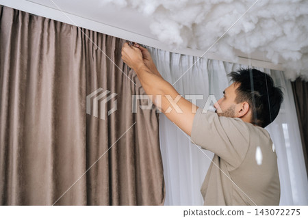 Low-angle view of man installing curtains on white rail, attaching fabric hooks to refresh a living room window during a home renovation, surrounded by playful cloud decorations. Low-angle view of man installing curtains on white rail, attaching fabric hooks to refresh a living room window during a home renovation, surrounded by playful cloud decorations. 143072275