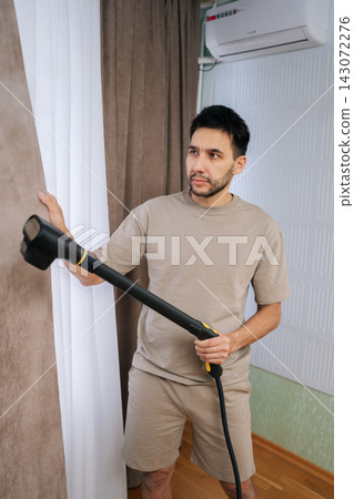 Vertical portrait of man steam cleaning curtains at home providing professional laundry service, achieving hygiene, freshness, performing housework and textile care. Concept of cleaning and household. 143072276
