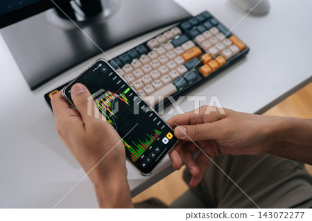 Close-up of trader analyzing real-time market data on smartphone, reviewing stock and crypto charts at desk with keyboard to monitor investments and make informed trading decisions. Close-up of trader analyzing real-time market data on smartphone, reviewing stock and crypto charts at desk with keyboard to monitor investments and make informed trading decisions. 143072277