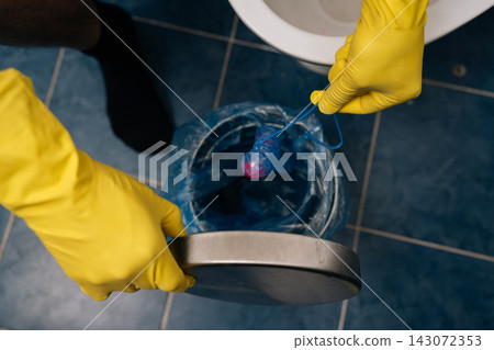 Human hands in yellow protective gloves dropping used toilet bowl cleaner block into lined rubbish bin, highlighting bathroom hygiene, waste disposal and household cleaning routine, close-up. Human hands in yellow protective gloves dropping used toilet bowl cleaner block into lined rubbish bin, highlighting bathroom hygiene, waste disposal and household cleaning routine, close-up. 143072353