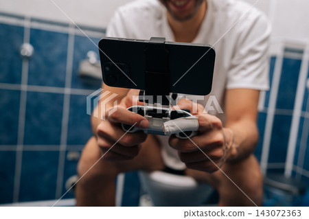 Unrecognizable relaxed young man holding smartphone with gamepad attachment and playing video games sitting on toilet, illustrating mobile gaming addiction, multitasking, privacy in bathroom. Unrecognizable relaxed young man holding smartphone with gamepad attachment and playing video games sitting on toilet, illustrating mobile gaming addiction, multitasking, privacy in bathroom. 143072363