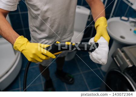 Closeup of cleaning company worker wearing yellow rubber gloves holding steam cleaner with white microfiber cloth, preparing to sanitize and deep clean the bathroom surfaces and tiles, close-up. Closeup of cleaning company worker wearing yellow rubber gloves holding steam cleaner with white microfiber cloth, preparing to sanitize and deep clean the bathroom surfaces and tiles, close-up. 143072364