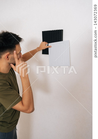 Vertical shot of pensive craftsman comparing different soundproofing acoustic panels and placing them on wall for noise reduction, improving room acoustics for sound recording or home studio. 143072369