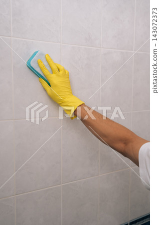 Close-up of unrecognizable person in yellow rubber glove using blue cleaning sponge to scrub and clean white ceramic tiles on bathroom wall, indicating domestic tasks and sanitation. Close-up of unrecognizable person in yellow rubber glove using blue cleaning sponge to scrub and clean white ceramic tiles on bathroom wall, indicating domestic tasks and sanitation. 143072373