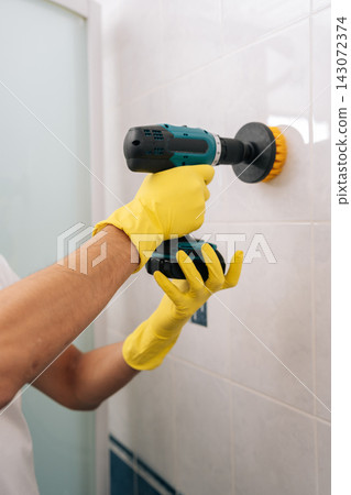 Vertical cropped shot of man using powerful electric drill scrubber to efficiently clean white bathroom tiles, speeding up routine housework with modern tools for meticulous, hygienic results. Vertical cropped shot of man using powerful electric drill scrubber to efficiently clean white bathroom tiles, speeding up routine housework with modern tools for meticulous, hygienic results. 143072374