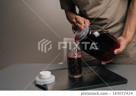 Detail cropped shot of unrecognizable person pouring fresh berry compote from large glass jar into drinking glass on grey table, preparing refreshing homemade fruit drink. Detail cropped shot of unrecognizable person pouring fresh berry compote from large glass jar into drinking glass on grey table, preparing refreshing homemade fruit drink. 143072424