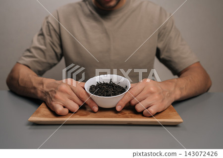 Cropped shot of man preparing tea leaves for brewing, holding bag of ingredients sitting at table, engaging in tranquil tea ceremony, mindful moment, close-up. Cropped shot of man preparing tea leaves for brewing, holding bag of ingredients sitting at table, engaging in tranquil tea ceremony, mindful moment, close-up. 143072426