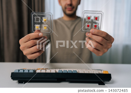 Man holding two packs of mechanical keyboard switches, comparing options for customizing or repairing a wireless mechanical keyboard on clean white desk, close-up cropped shot. Man holding two packs of mechanical keyboard switches, comparing options for customizing or repairing a wireless mechanical keyboard on clean white desk, close-up cropped shot. 143072485