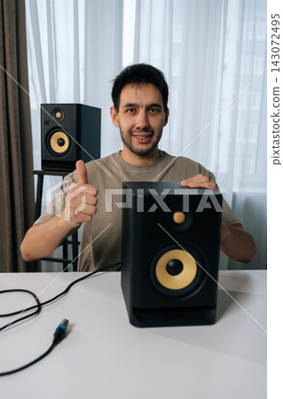 Vertical shot of man smiling looking camera and giving thumbs up gesture, connecting studio monitor speaker for home recording or music production setup, expressing satisfaction with audio equipment. Vertical shot of man smiling looking camera and giving thumbs up gesture, connecting studio monitor speaker for home recording or music production setup, expressing satisfaction with audio equipment. 143072495