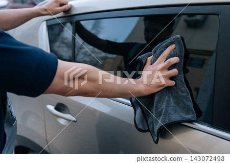 Person wiping car side window with gray microfiber cloth, detailing vehicle by hand for spotless glass and professional auto care service in workshop setting.. 143072498