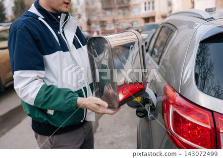 Man holding metal jerrycan, pouring gasoline into cars tank on city street, highlighting emergency refueling, fuel shortage and transportation challenges amid rising prices.. Man holding metal jerrycan, pouring gasoline into cars tank on city street, highlighting emergency refueling, fuel shortage and transportation challenges amid rising prices.. 143072499
