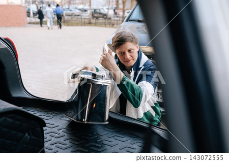 Young man in retro jacket puts shiny metal jerrycan into car trunk. View from inside vehicle showing preparation for road trip or fuel emergency. Urban parking lot background with natural light. Young man in retro jacket puts shiny metal jerrycan into car trunk. View from inside vehicle showing preparation for road trip or fuel emergency. Urban parking lot background with natural light. 143072555